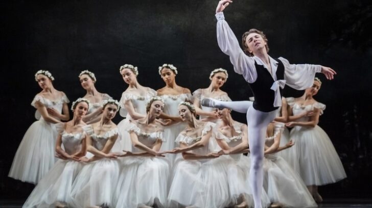 Male ballet dancer in black tunic dances in front of large group of female ballet dancers in white dresses