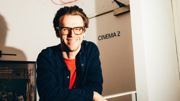 person smiling with square glasses leans on railing before sign saying cinema 2