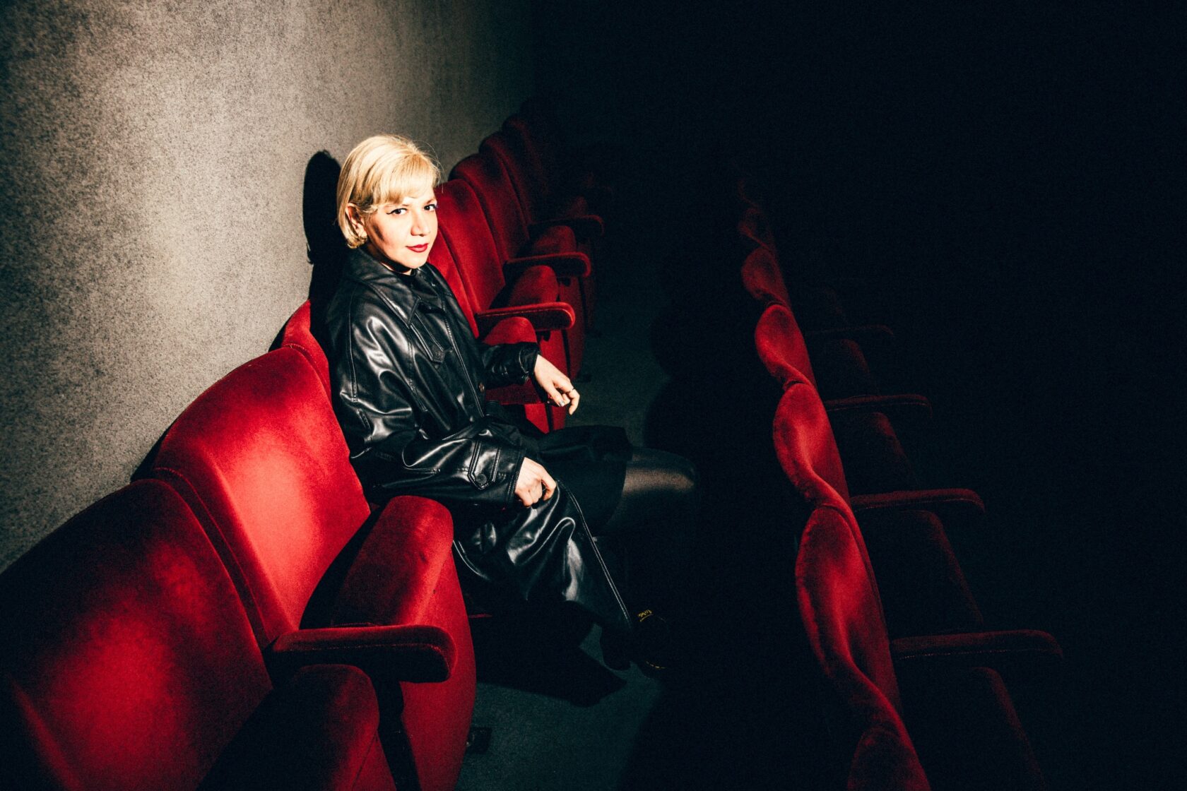 person with short blonde hair in leather jacket sitting on red velvet cinema seats