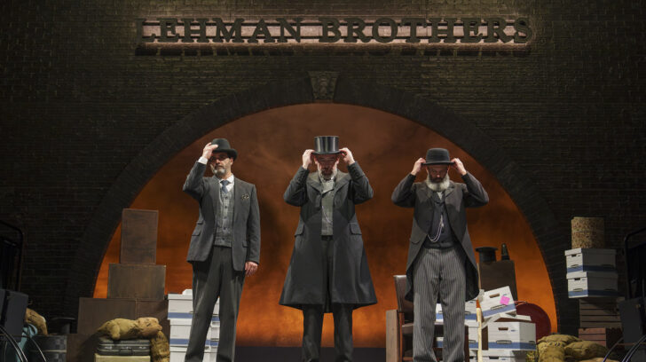 Three men on stage in suits, lifting top hat from head